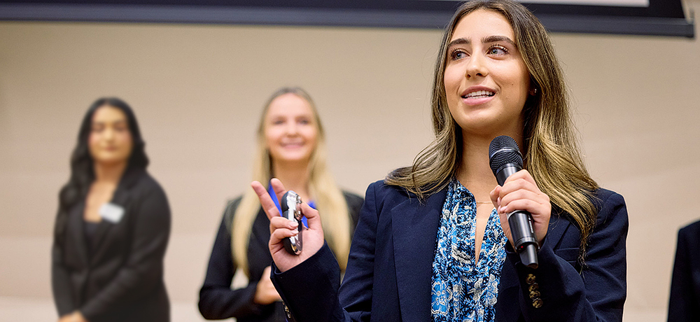 Section Image: Female student presenting with microphone in one hand and clicker in the other with members of the team behind 